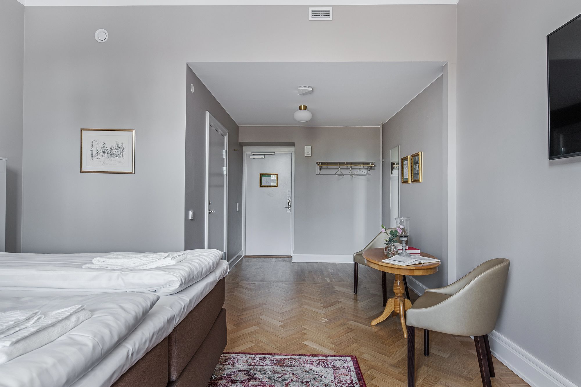 Ein Hotelzimmer mit Bett, Schreibtisch mit Stuhl, Spiegel und einem kleinen Tisch mit Vase.