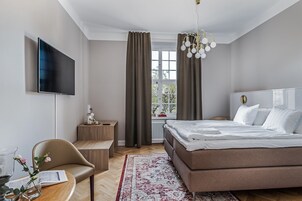 Ett modernt hotellrum med en stor säng, en tv som är monterad på väggen, ett litet träbord, en stol och ett fönster med gardiner.