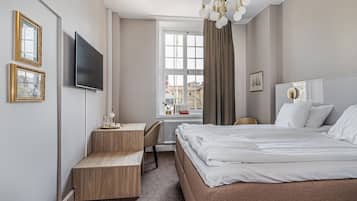 A hotel room with a bed, a desk, a chair, a TV, a window with curtains, and a radiator.
