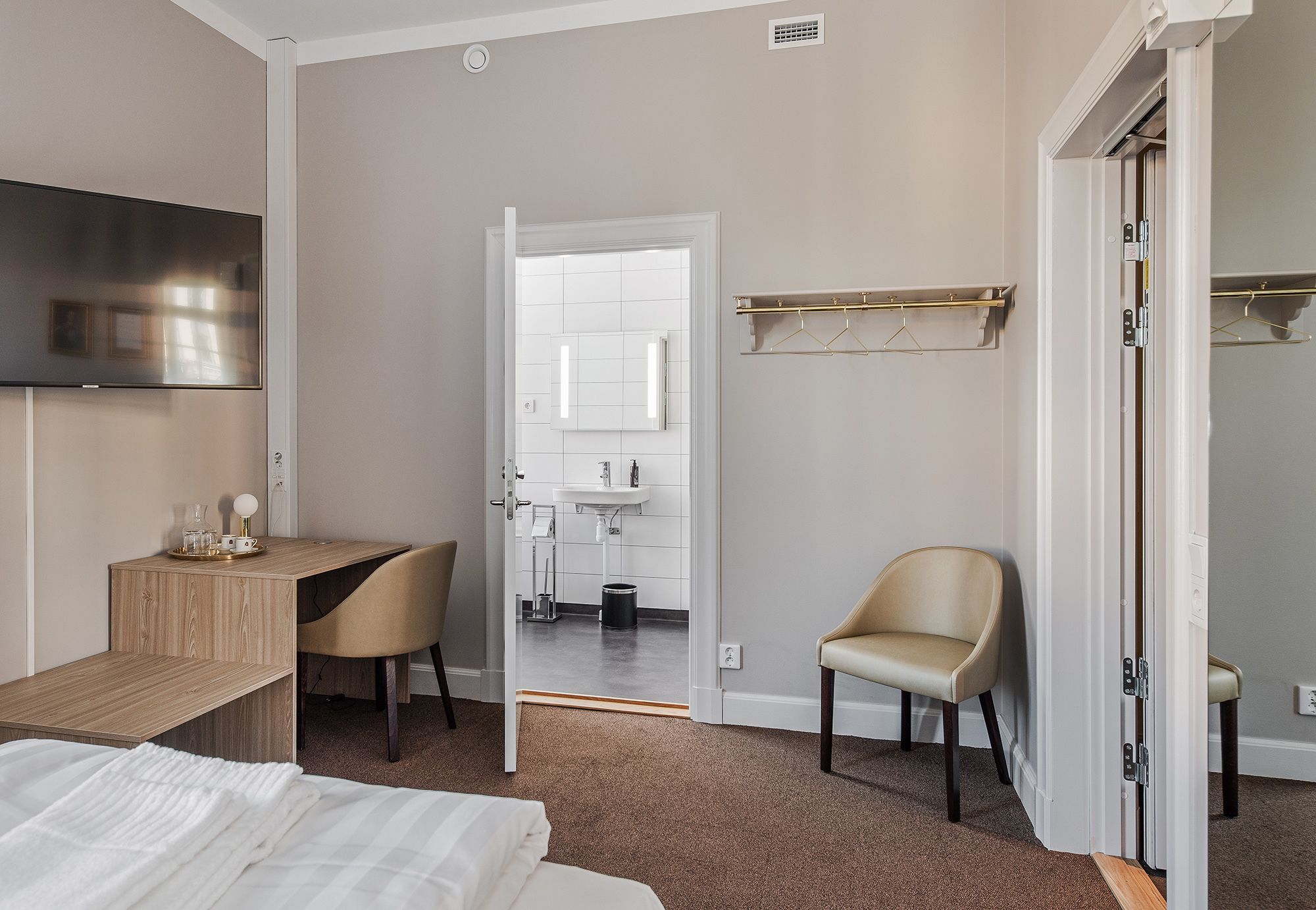 Ein Hotelzimmer mit Bett, Schreibtisch, Stuhl, Fernseher sowie einem durch eine offene Tür einsehbarer Badezimmer und einem Kleiderständer.