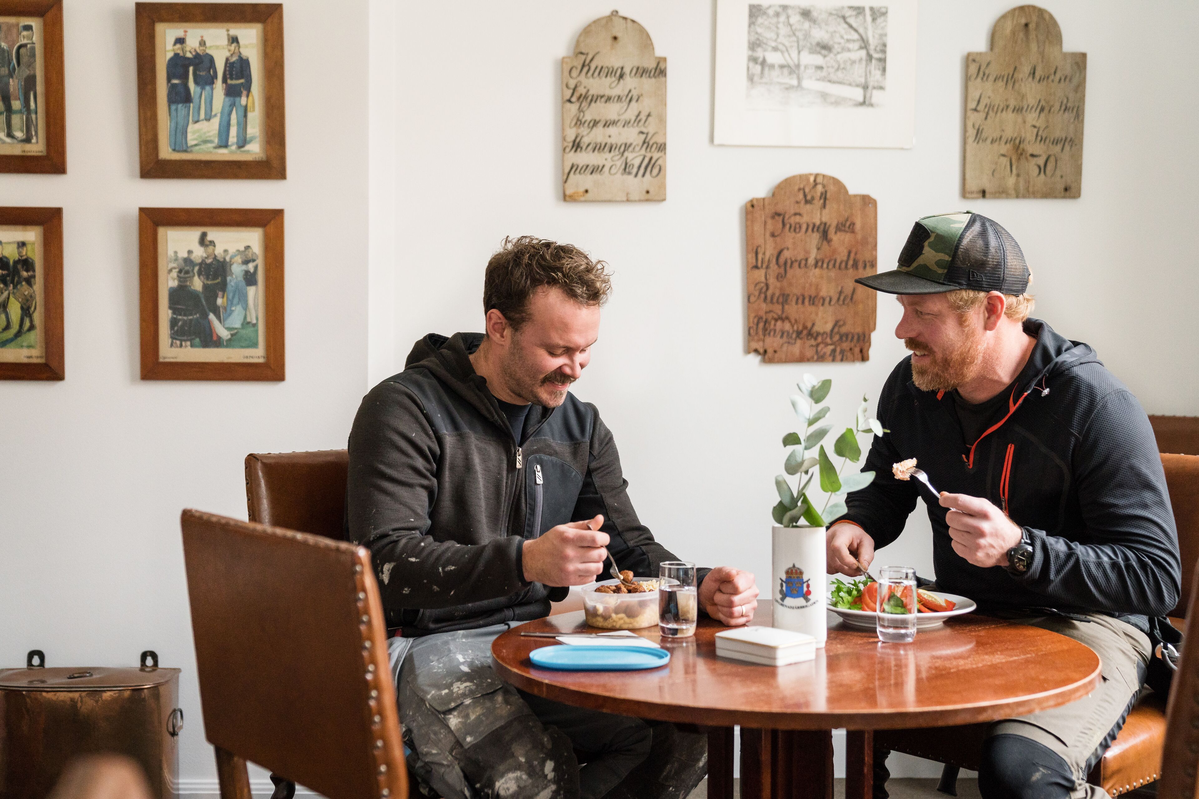 Two people dining at a round wooden table with framed pictures on the wall behind them.
