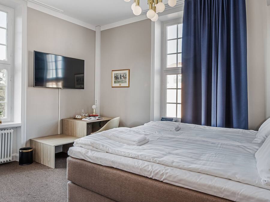 A bedroom with a bed, a desk, a chair, a radiator, a TV, and a window with curtains.