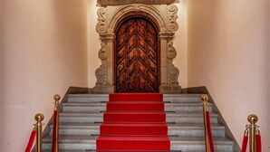 Eine prächtige Treppe mit rotem Teppich, die zu einer verzierten Holztür führt, gesäumt von roten Samtseilen und goldenen Ständern.