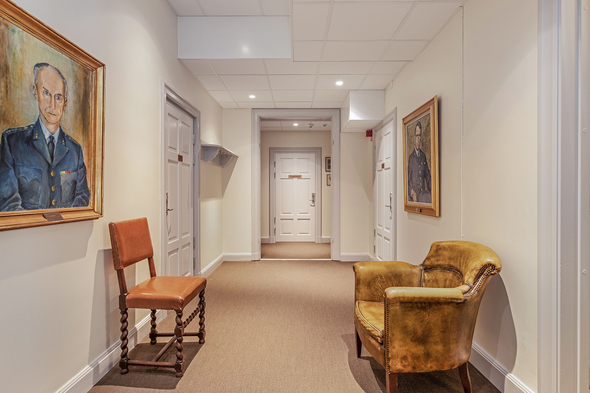 Un couloir avec deux chaises, une porte portant l’inscription « Direction » et un tableau accroché au mur.