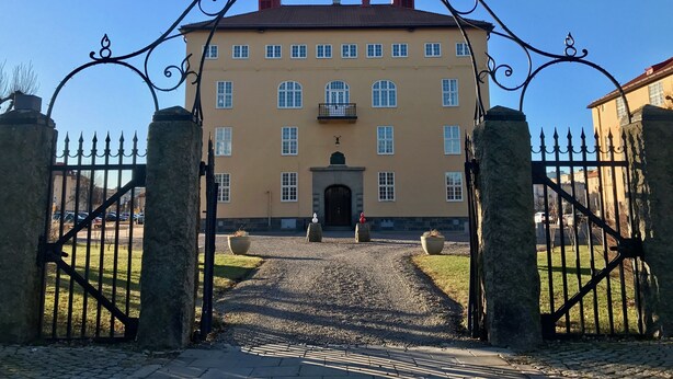 Ein großes Gebäude mit einer symmetrischen Fassade, einem zentralen Eingang mit einem dekorativen Bogen und einer Schotterauffahrt zum Eingang.