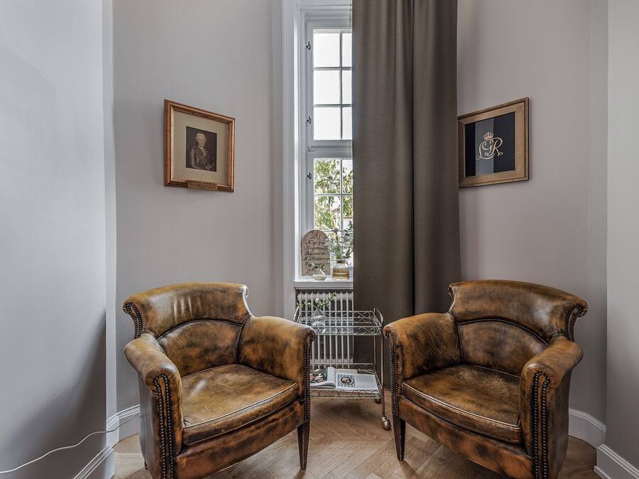 Une pièce comprenant deux fauteuils en cuir, un radiateur, une fenêtre avec des rideaux et des tableaux encadrés accrochés aux murs.