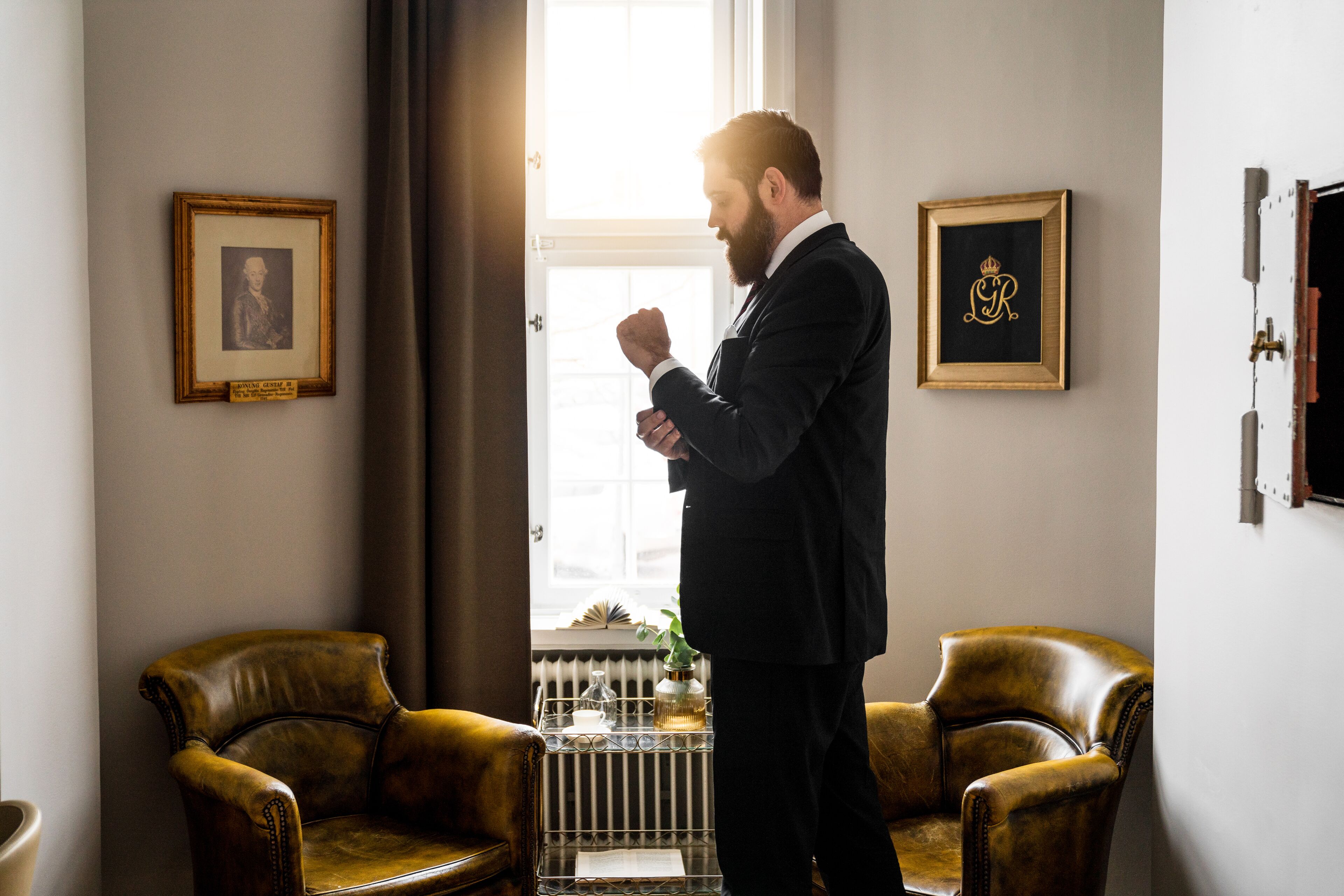 Un homme en costume se tient debout dans une pièce où se trouvent deux fauteuils en cuir, un radiateur et des tableaux encadrés accrochés au mur.