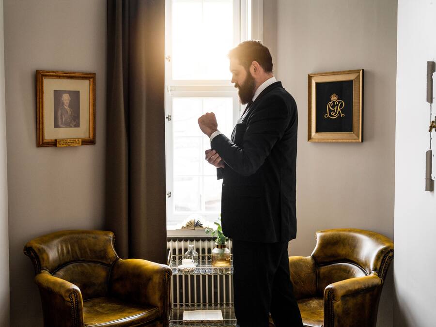 Un homme en costume se tient debout dans une pièce où se trouvent deux fauteuils en cuir, un radiateur et des tableaux encadrés accrochés au mur.