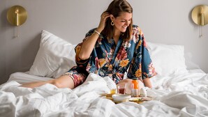 Eine Person sitzt auf einem Bett mit Tablett und Frühstück, einer Wandleuchte und gerahmten Bildern.