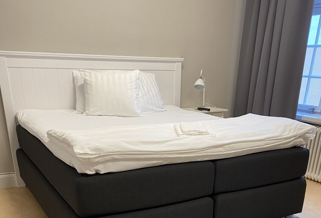 A neatly made bed with white linens and a black headboard in a room with a window and a nightstand with a lamp.