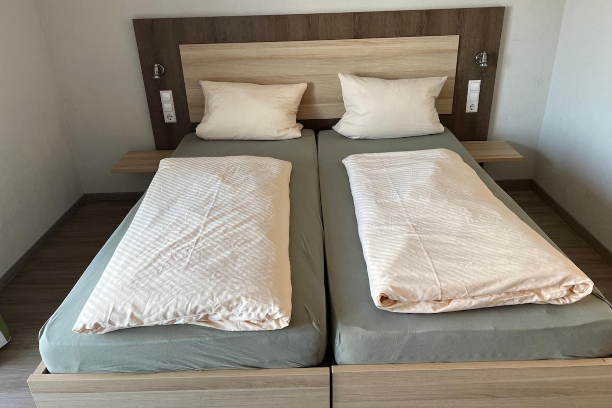 Uma cama de casal com duas almofadas e duas cobertas, cabeceira de madeira e mesas de cabeceira com abajures.