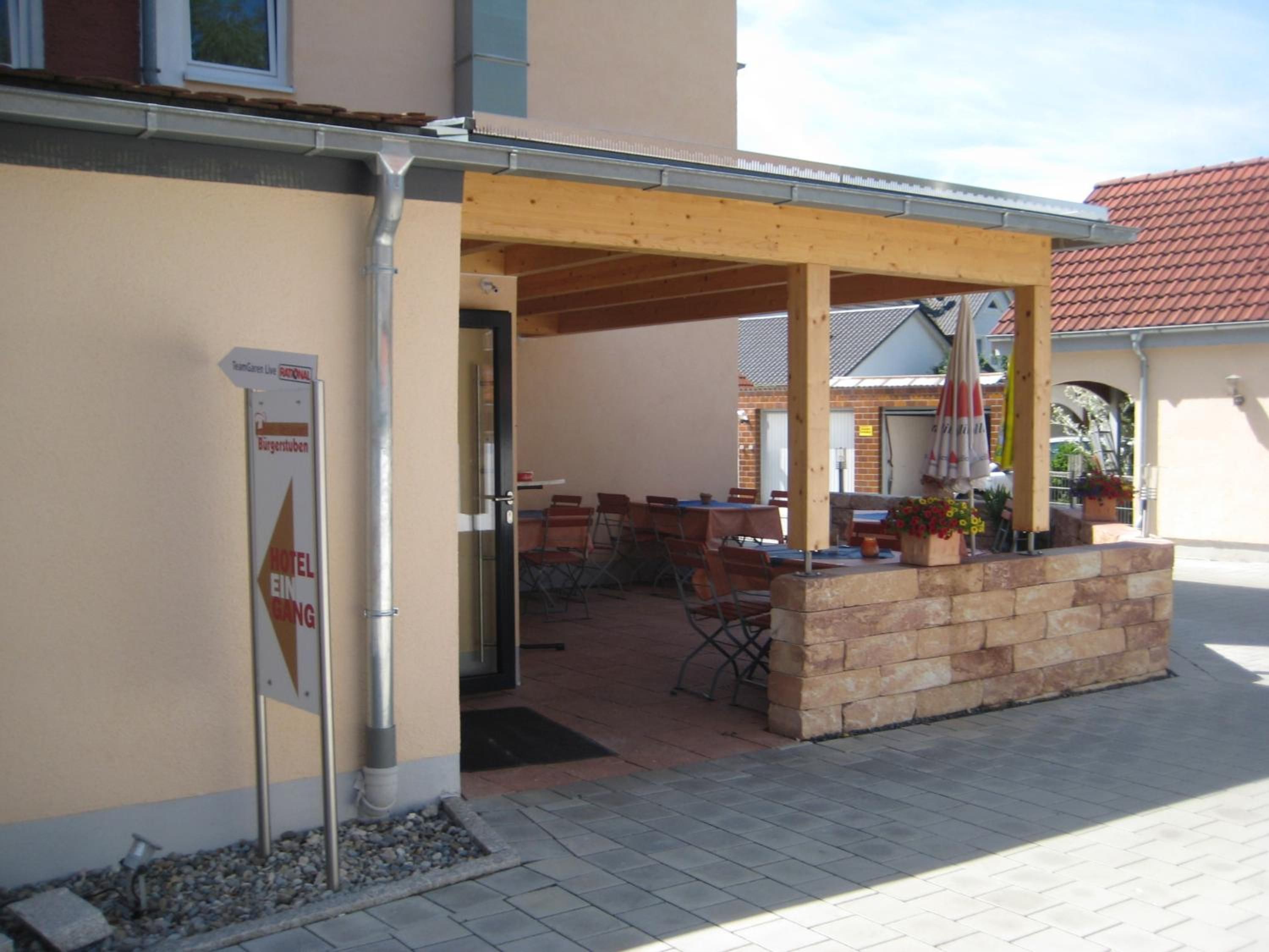Pátio externo com mesas e cadeiras, pergolado de madeira e placa indicando um hotel.