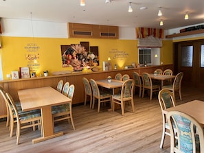 A restaurant interior with wooden tables and chairs, a yellow wall with text and a fruit display, and pendant lighting.