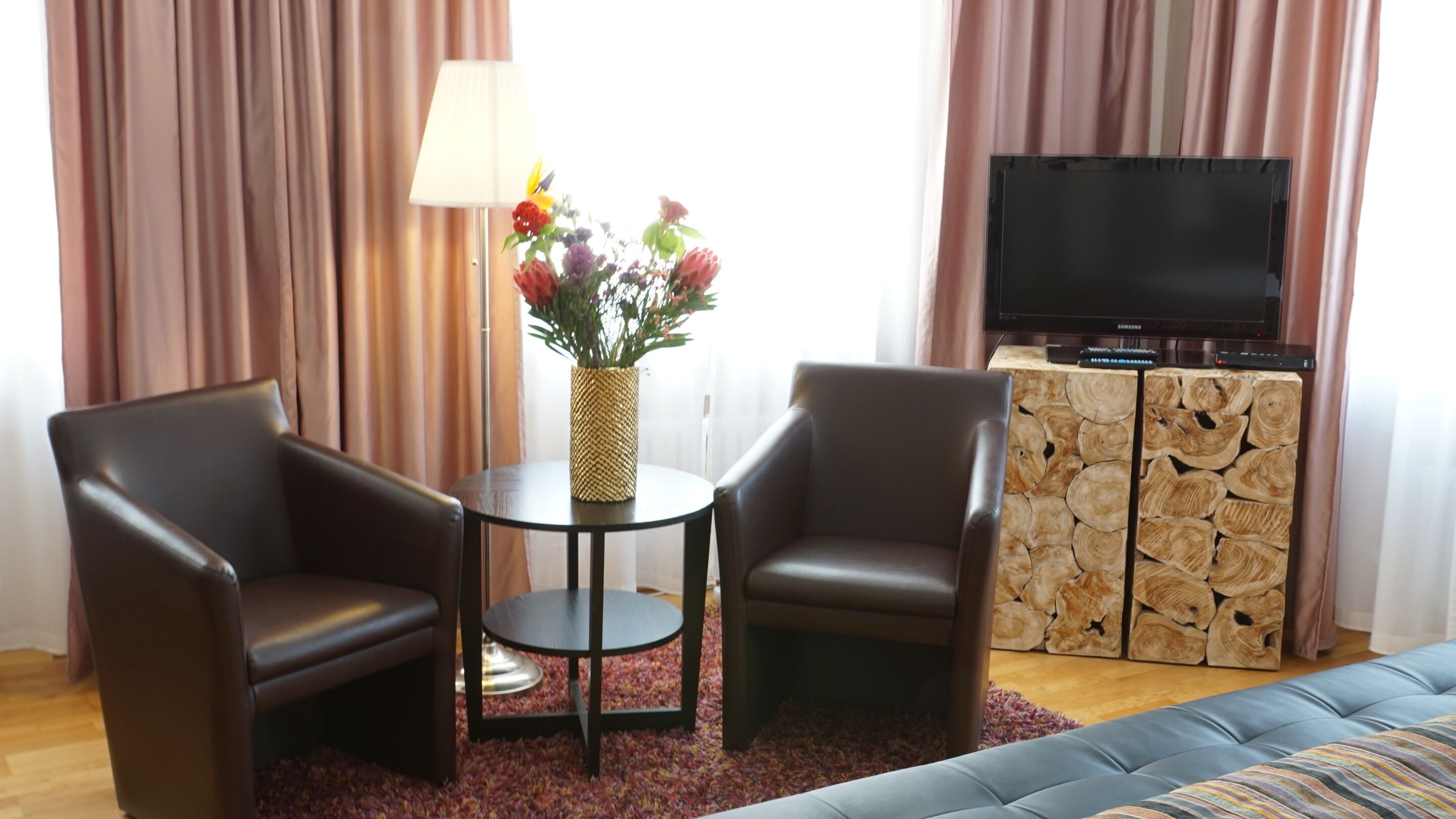 A room with two leather armchairs, a small round table with a vase of flowers, a television on a wooden cabinet, and a sofa.