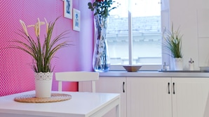 Una cocina con armarios blancos, una mesa blanca y una pared rosa con lunares. Hay plantas en macetas y un florero con flores en la encimera.