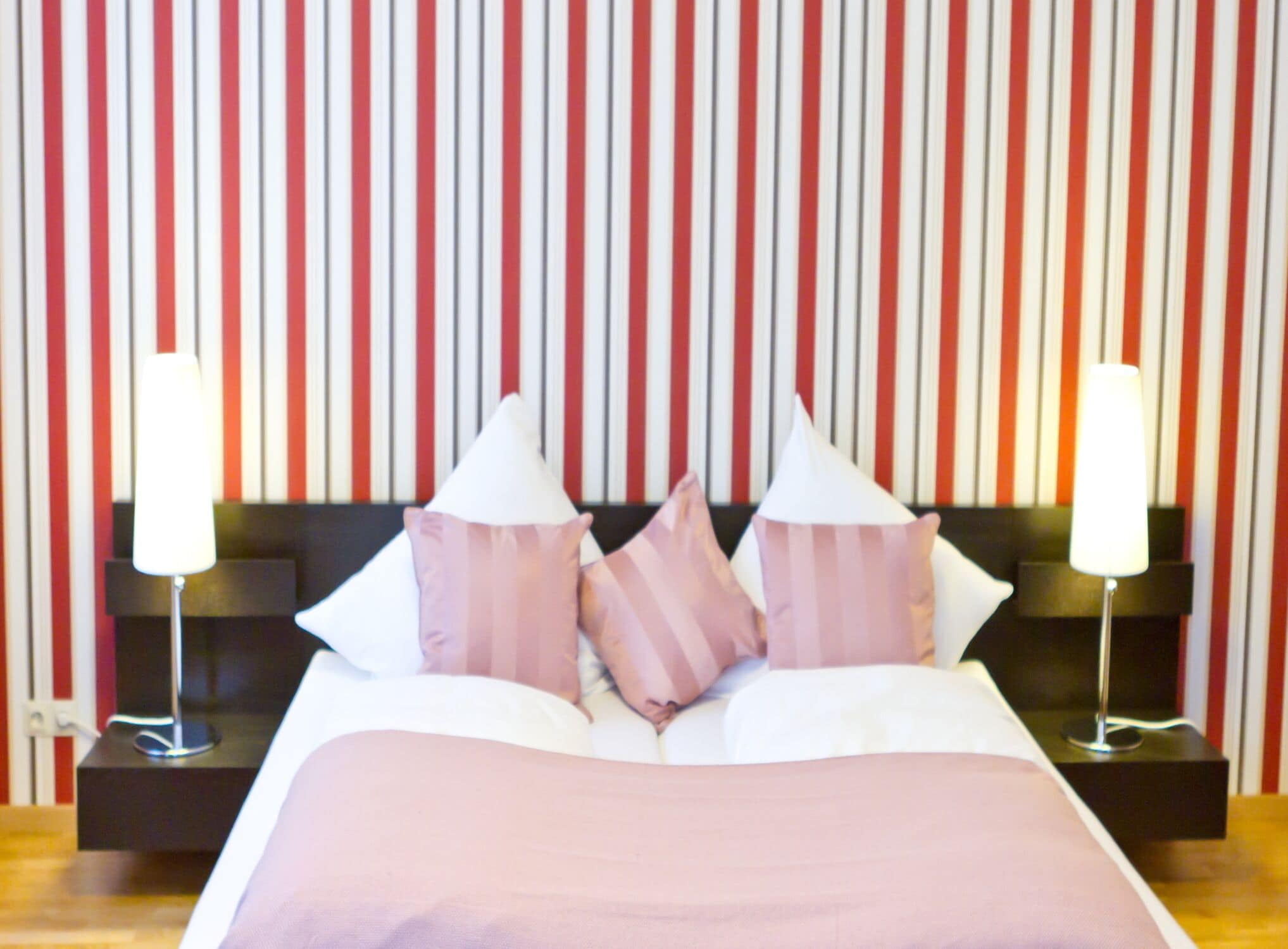Une chambre d’hôtel avec un mur à rayures, un lit avec une literie rose et blanche, et deux lampes de chevet.