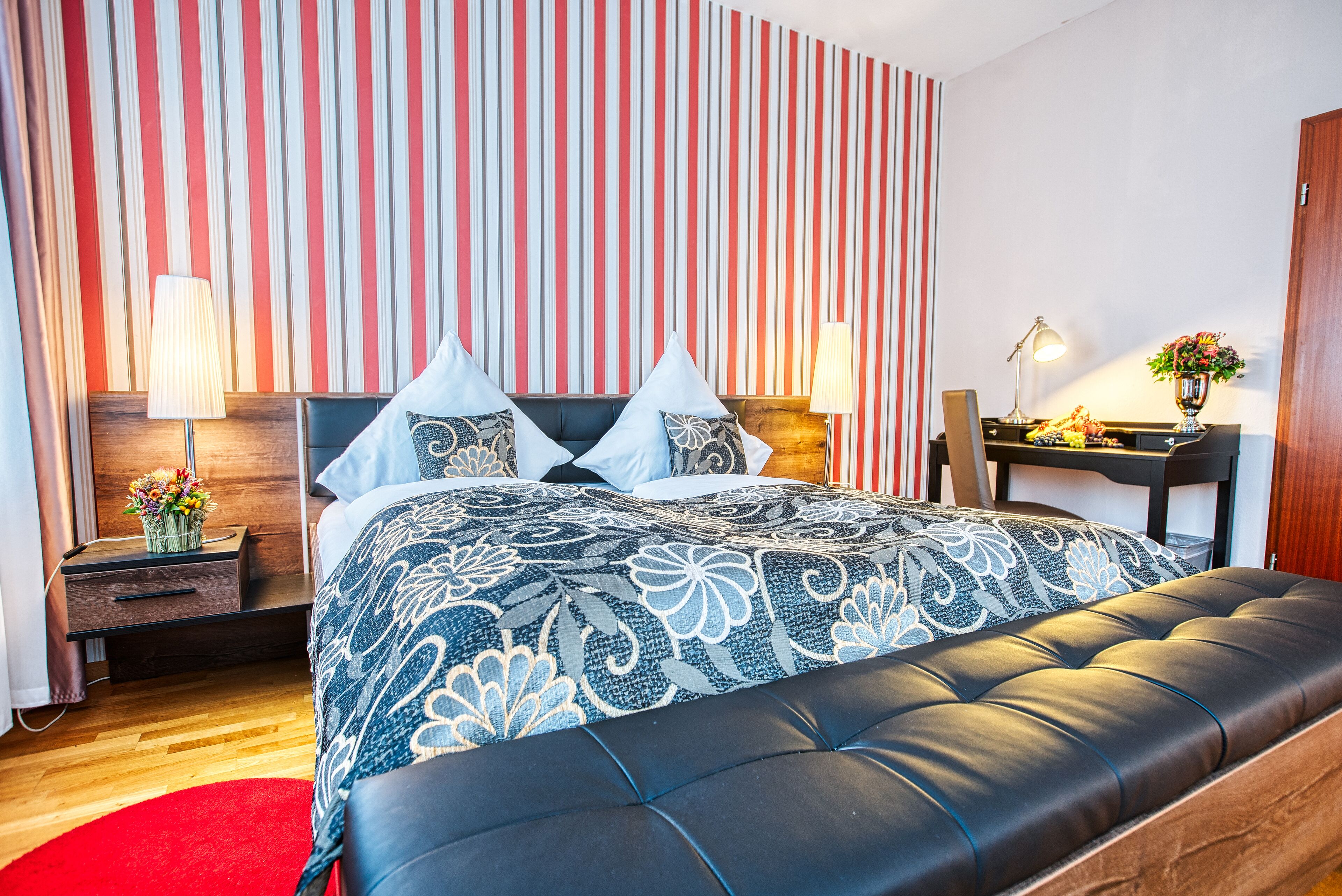 Ein modernes Schlafzimmer mit gestreifter Tapete, einem großen Bett mit einem gemusterten Überwurf, einem Nachttisch aus Holz, einem Schreibtisch mit Lampe und einem roten Teppich.
