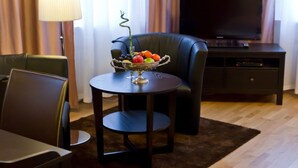 Habitación de hotel con un sofá de cuero oscuro, una mesita redonda con frutero, una lámpara y un televisor sobre un mueble de madera.