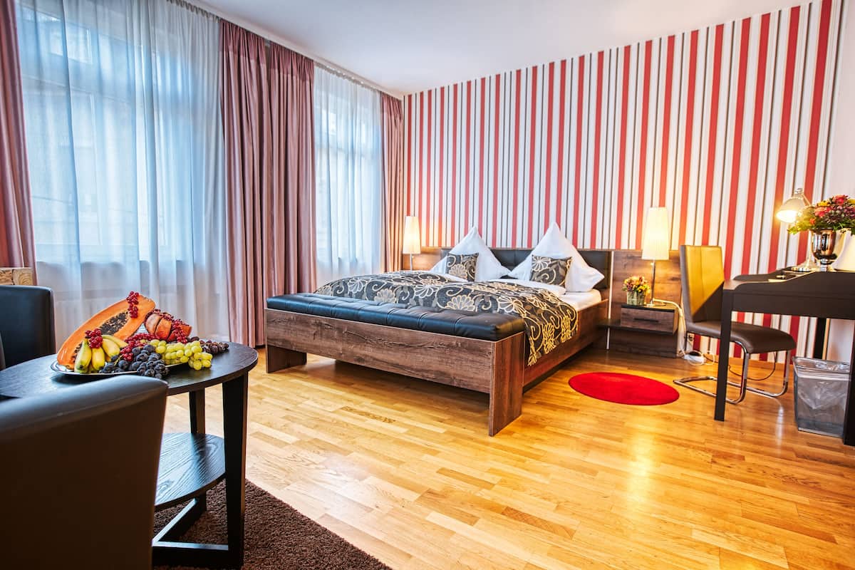 A modern hotel room with a wooden bed, striped wallpaper, a dining table with fruit, and a red rug.