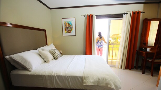 A hotel room with a large bed, a desk, and a chair. There is a window with curtains and a painting on the wall. - Luxury Hotel & Apts (Georgetown)