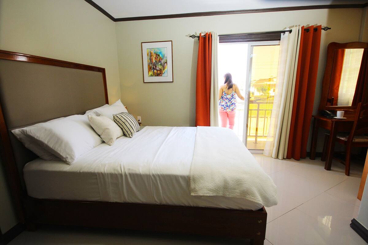 A hotel room with a large bed, a desk, and a chair. There is a window with curtains and a painting on the wall.
