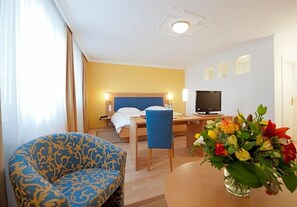 Una habitación de hotel con cama, escritorio con televisor, una silla azul y un florero con flores.