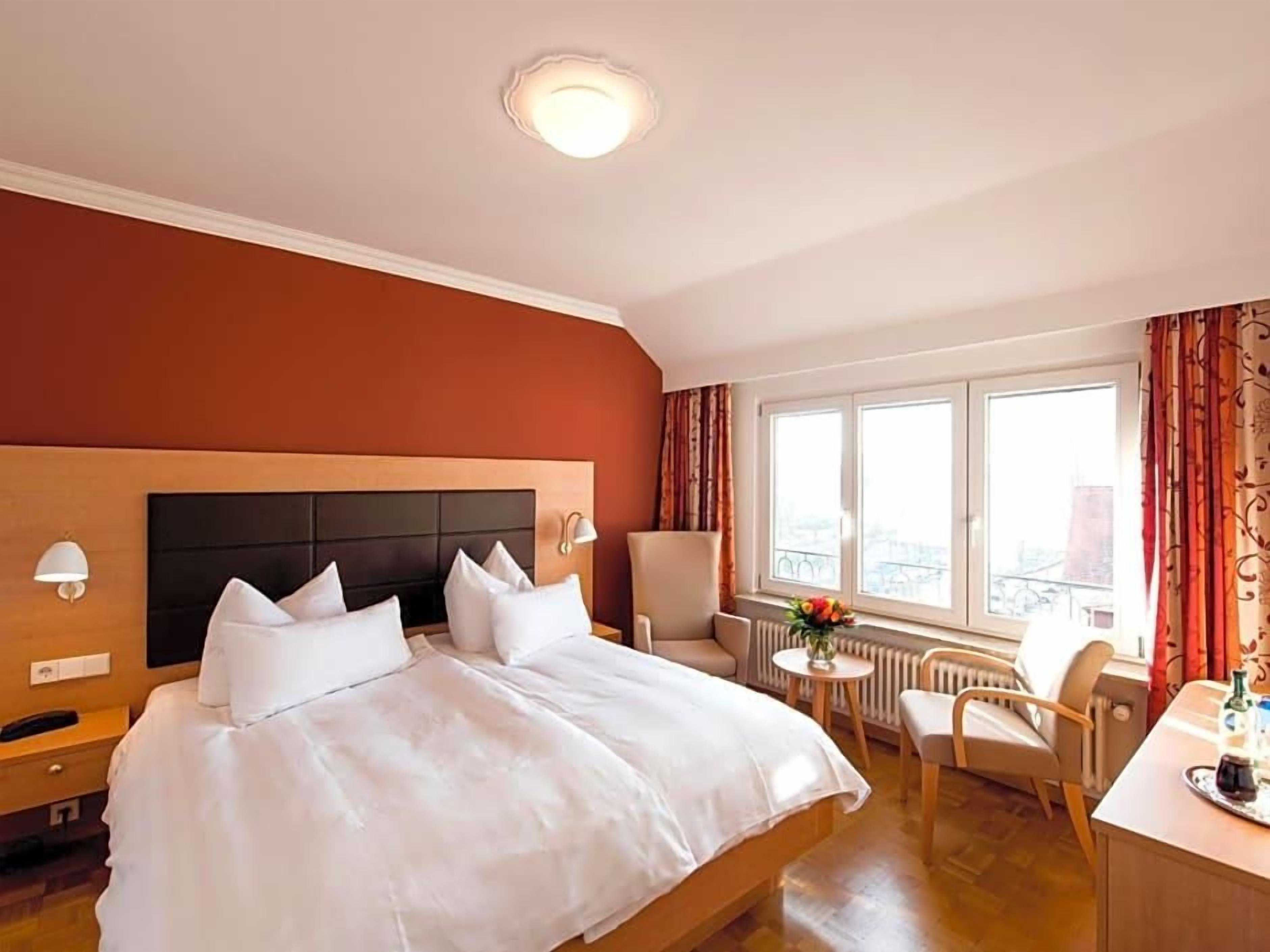 Ein Hotelzimmer mit einem großen Bett, einem Schreibtisch, einem Stuhl, einem kleinen Tisch und einem Fenster mit Vorhängen.