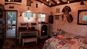 Interior rústico de una cabaña con pared de madera, chimenea, cama con colcha y cabeza de venado montada en la pared, además de una puerta que da al exterior.