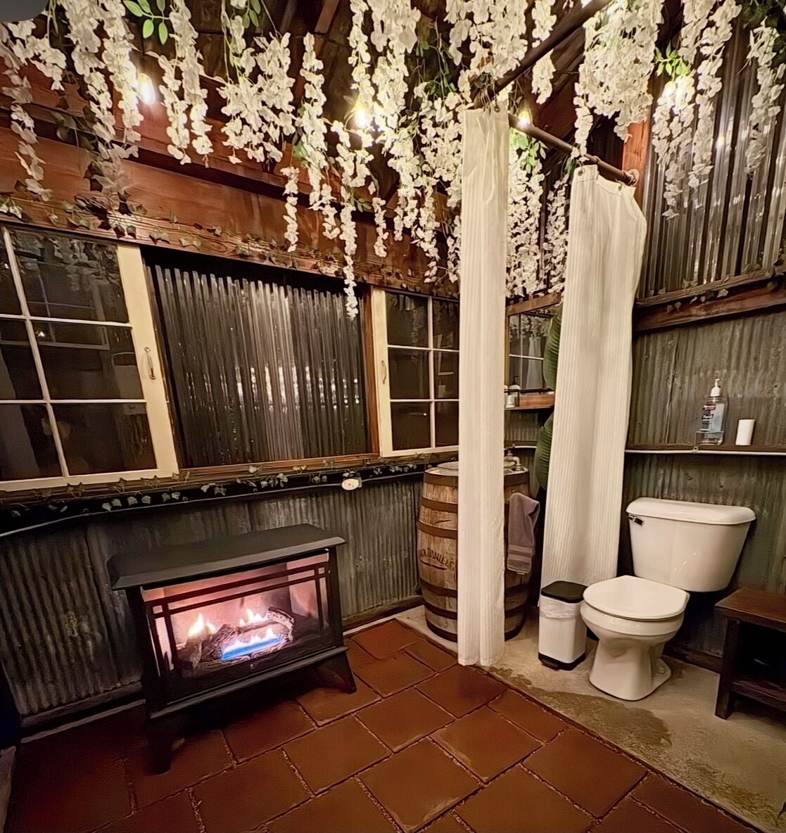 A rustic bathroom with a wood-paneled wall, a stone fireplace, a toilet, and a shower curtain.