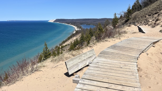 A wooden boardwalk leading to a sandy beach with a view of the ocean and distant hills.