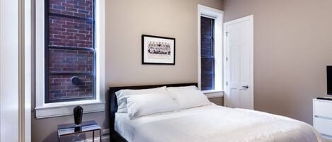 Ein Schlafzimmer mit einem Bett, einem Fenster mit Blick auf eine Backsteinmauer, einem Deckenventilator und einem gerahmten Bild an der Wand.
