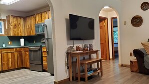 Una cocina moderna con armarios de madera, un refrigerador y un microondas. Hay un televisor de pantalla plana montado en la pared, una mesa de madera con un teléfono y un sofá con funda acolchada.