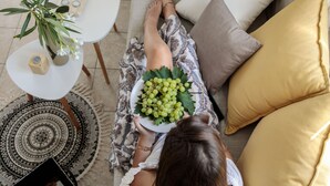 Eine Person sitzt auf einem Sofa mit einer Schüssel grünen Trauben, einer Topfpflanze und einem weißen Tisch mit Kerze und einer Kiste.