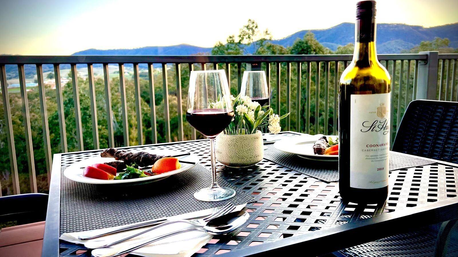 A table set for a meal with a bottle of wine, a glass, and a plate of food on a balcony overlooking a mountainous landscape.