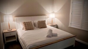 Een net opgemaakt bed met een houten hoofdeinde, twee bedlampen en een raam met zonwering.