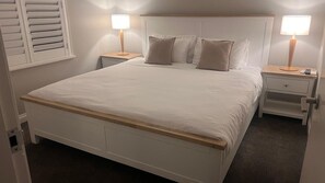 Una cama bien hecha con cabecera de madera, dos mesitas de noche con lámparas y una ventana con persianas.