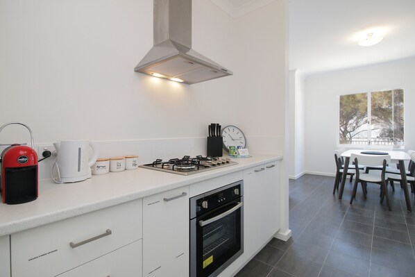 A modern kitchen with a gas stove, oven, and coffee maker. There is a dining area with a table and chairs. - Estuary Waterfront - Upstairs with a small Balcony (Falcon)