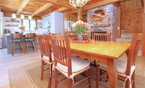 Une table à manger en bois, entourée de chaises, une cheminée et, au fond, un coin cuisine.