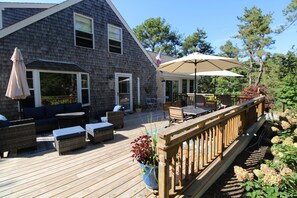 A wooden deck with outdoor furniture, a covered patio area, and a view of a house with a dark shingled exterior. - Lovely Post & Beam Near Indian Neck (1224) (Wellfleet)