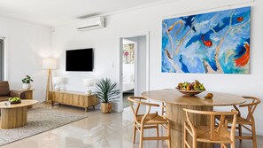 Una sala de estar moderna con un televisor de pantalla plana, una zona de comedor con una mesa redonda y sillas de mimbre, y un cuadro abstracto grande en la pared.