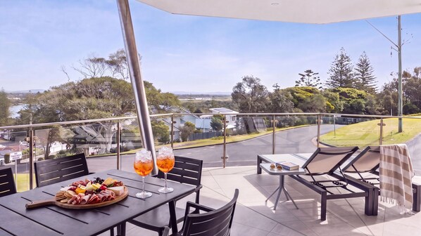 An outdoor patio with a table set for dining, two drinks, and a view of the surrounding landscape. - Pilotage 1 - Elevated Yamba Escape with River, Ocean & Sunset Views - Holiday Yamba (YAMBA)