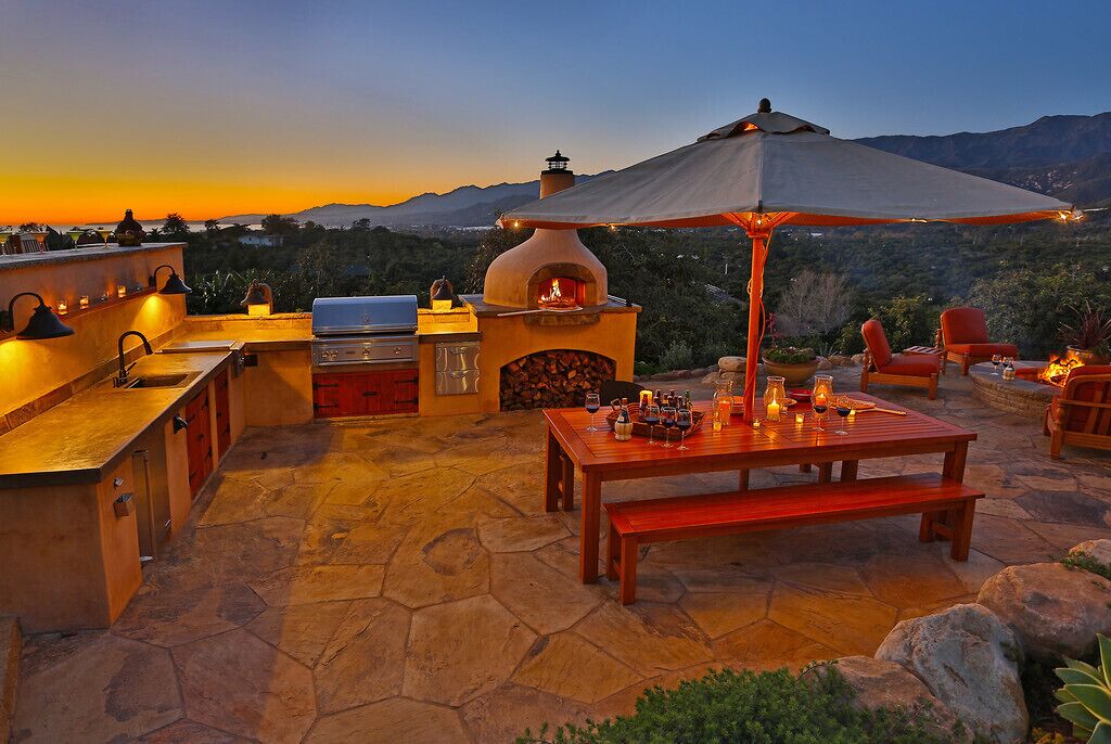 An outdoor patio with a stone floor, a pizza oven, a grill, and a table set for dining with candles and drinks.