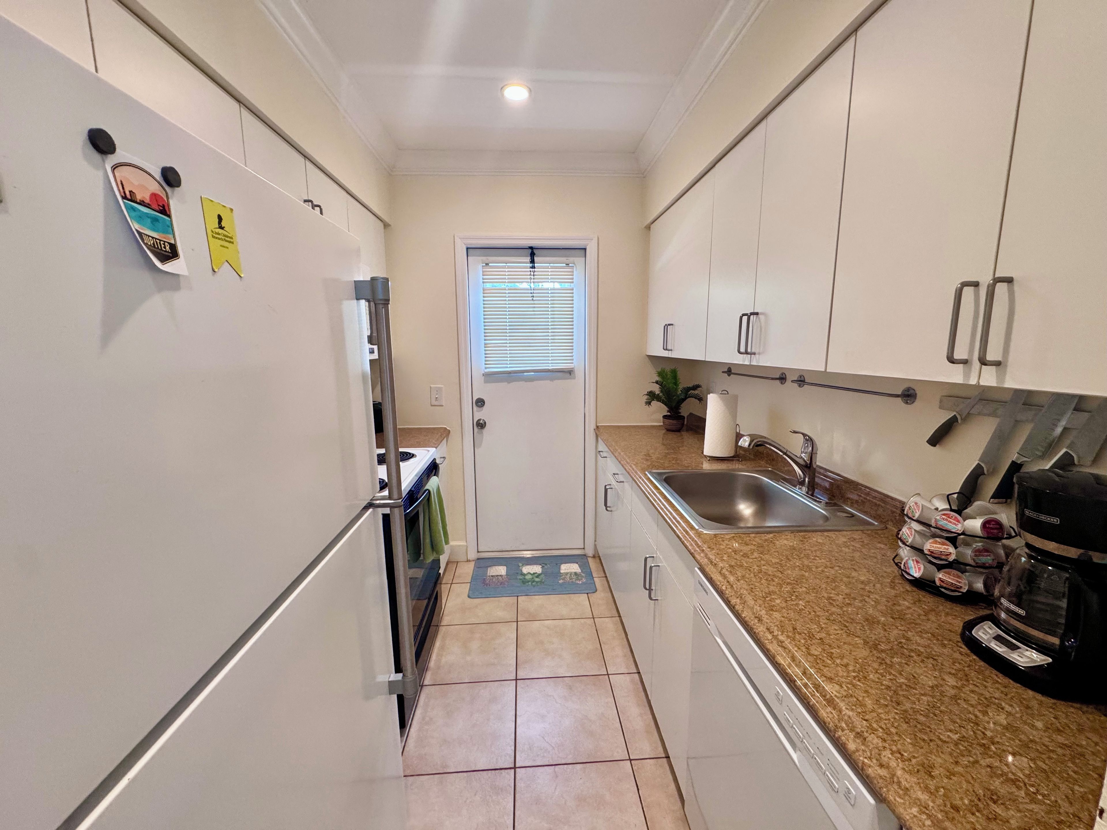A narrow kitchen with white cabinets, a stainless steel sink, and a coffee maker.