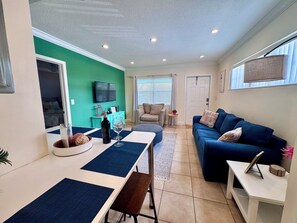 Ein modernes Wohnzimmer mit einem blauen Sofa, einem Esstisch mit einer Bank, einem an der Wand montierten Fernseher und einer Tür zu einem anderen Raum.