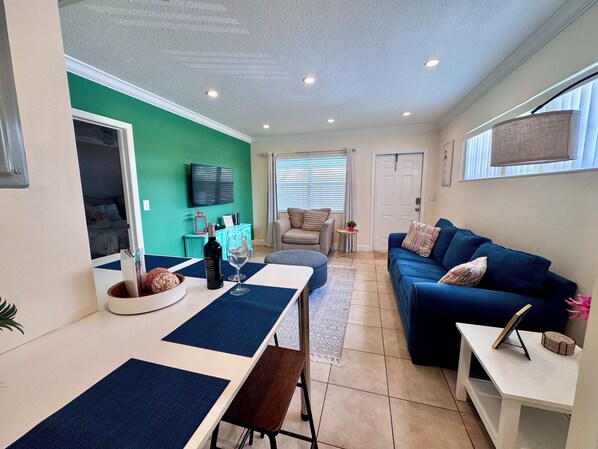 A modern living room with a blue sofa, a dining table with a bench, a television mounted on the wall, and a door leading to another room. - Two Bedroom Retreat Near Deerfield Beach Pier (Deerfield Beach)