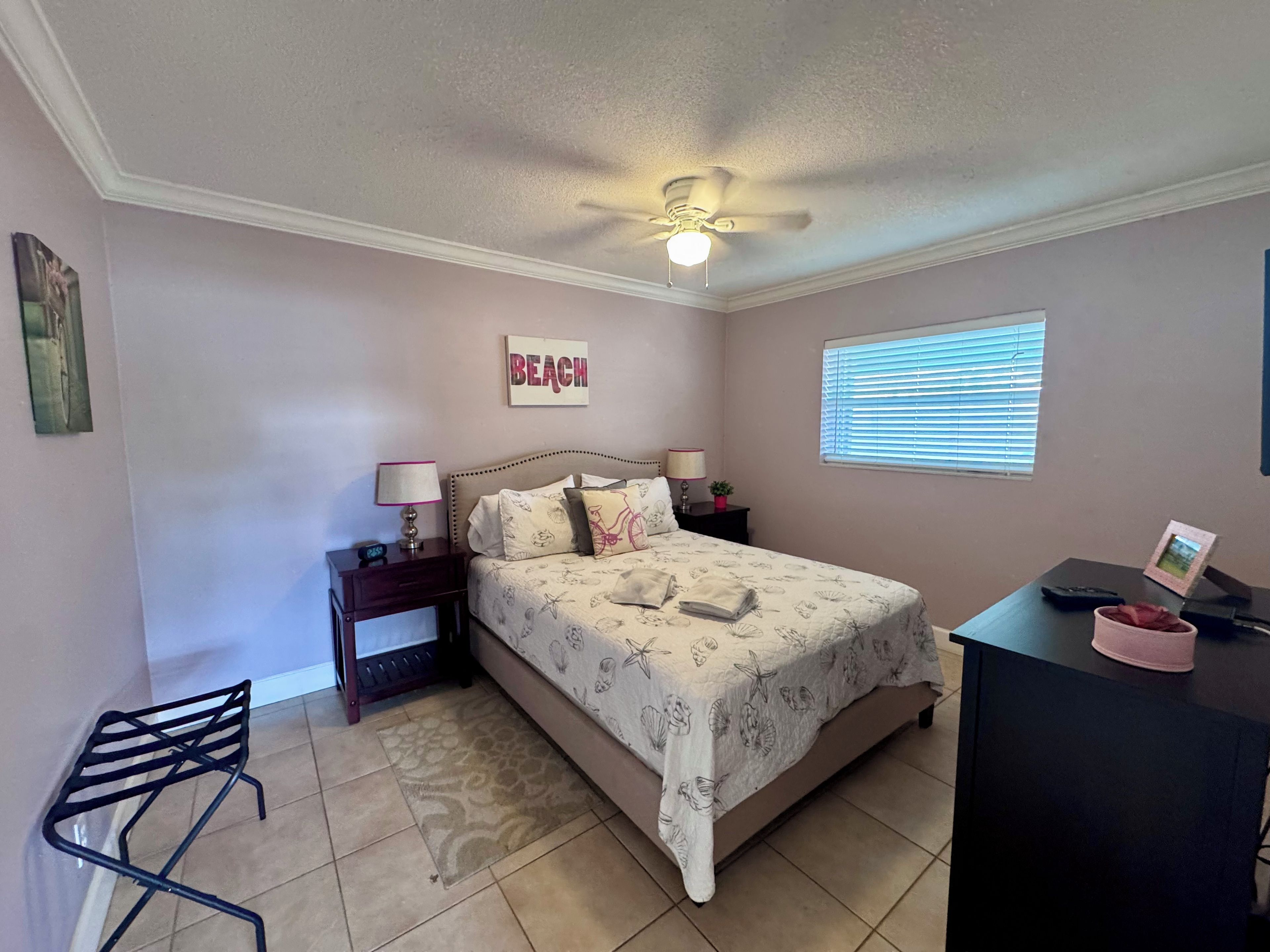 A bedroom with a bed, nightstands, a desk, a chair, a ceiling fan, a window with blinds, and a wall-mounted picture.