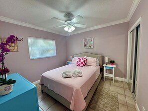 Ein Schlafzimmer mit einem Bett, einem Nachttisch, einem Deckenventilator, einem Fenster mit Jalousien und einem an der Wand hängenden Bild.