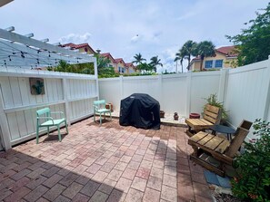 Eine Terrasse mit weißem Zaun, einer überdachten Grillstation, einer Holzbank und einem hellblauen Stuhl.