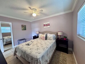A bedroom with a bed, two bedside tables, a ceiling fan, a window with blinds, and a door leading to another room.