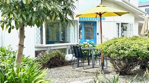 A patio with a table and chairs, a yellow umbrella, and a house with a blue door and windows.